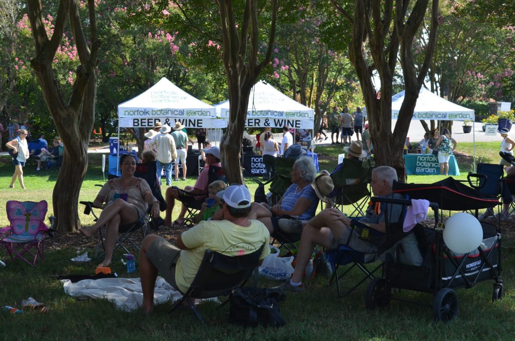 guests on lawn chairs sit outside under crepe myrtles with vendor tents in the background.